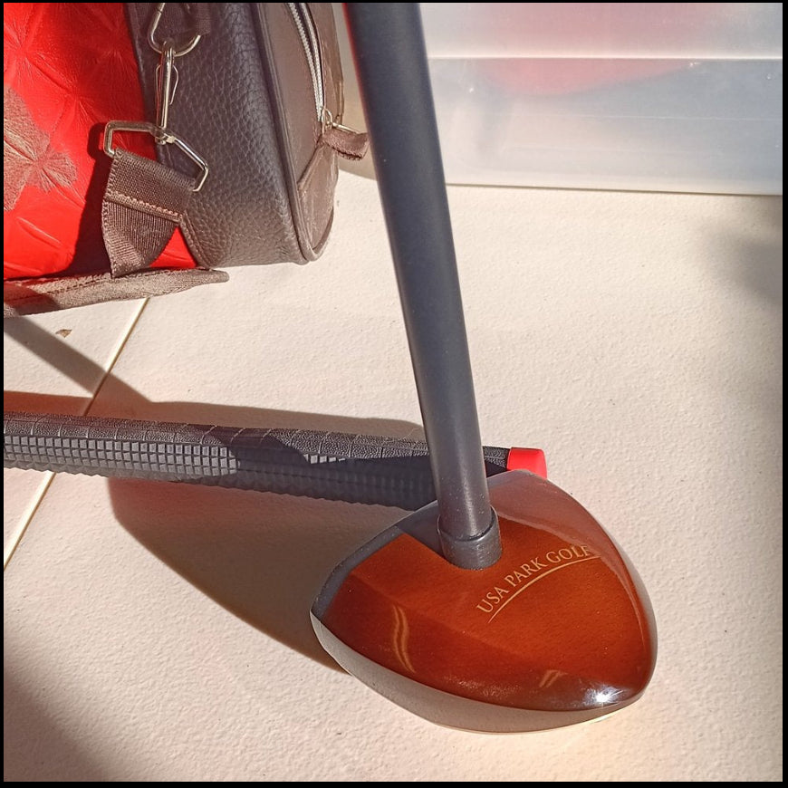 Close-up view of a USA Park Golf club head resting on a light beige surface. The club head is dark reddish-brown wood with the words "USA PARK GOLF" engraved in gold lettering.  A dark grey shaft extends upward, and a portion of a black grip is visible in the background. A brown leather golf bag is visible in the upper left corner.