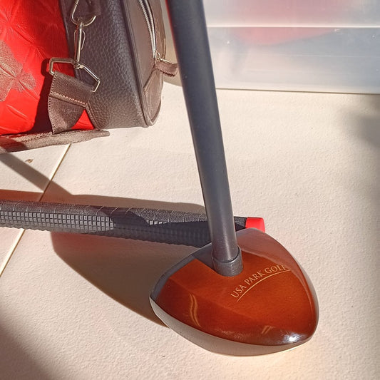 Close-up view of a USA Park Golf club head resting on a light beige surface. The club head is dark reddish-brown wood with the words "USA PARK GOLF" engraved in gold lettering.  A dark grey shaft extends upward, and a portion of a black grip is visible in the background. A brown leather golf bag is visible in the upper left corner.